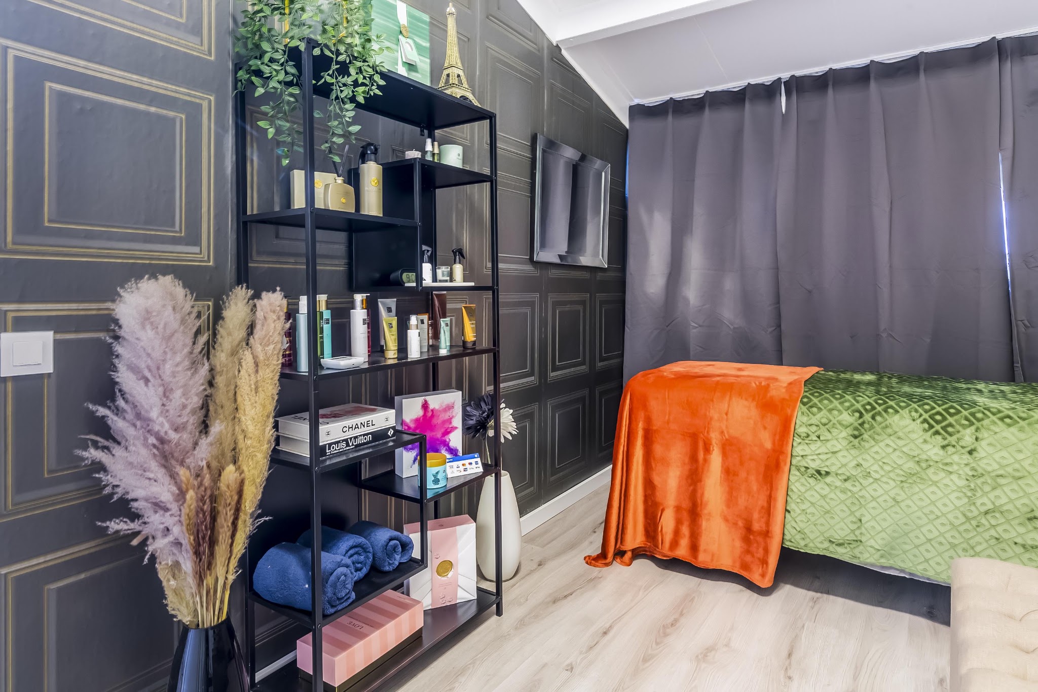 Behandelbed met oranje plaid in de salon, met een zwarte kast vol producten en pampasgras